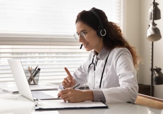 A person sitting at a laptop with a headset and a white physicians coat.