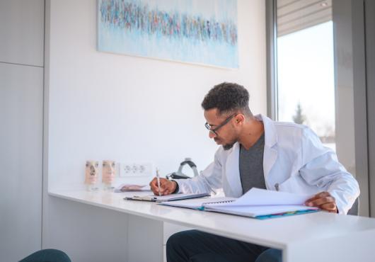 A doctor in a white coat sits at a desk writing. 