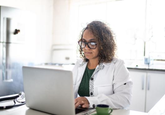 A doctor with glasses and a white coat types on a laptop with a stethoscope to the left of them and a cup of tea to the right.