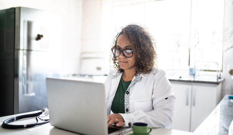 A doctor with glasses and a white coat types on a laptop with a stethoscope to the left of them and a cup of tea to the right.